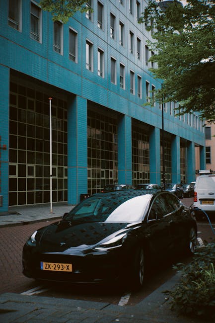 A sleek black electric car with a glossy finish and modern design is parked in a designated parking space in front of a large urban building with a bright blue tiled facade. The building features multiple windows arranged in a grid pattern, with some open and others closed, reflecting the surrounding environment. To the right, additional vehicles, including a white van, are parked along the street. The scene is set during daylight hours, with sunlight casting soft shadows, and lush green trees frame the top of the image, adding a touch of nature to the urban landscape. This setting highlights a clean, organized parking area, with the electric vehicle and surrounding architecture emphasizing contemporary urban design and climate-conscious transportation, all under the expertise of Gardeners Holland Park in maintaining aesthetic and functional outdoor spaces.