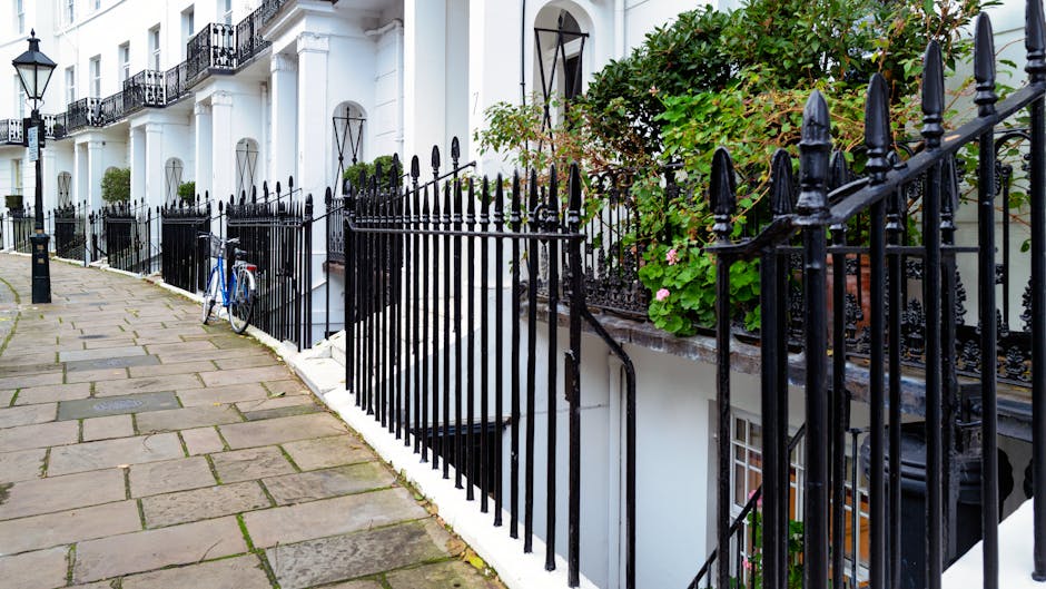 A cozy London garden scene featuring a variety of lush green plants on a brick patio enclosed by brick and wooden walls. There are potted plants with ferns and small shrubs, some with textured foliage, placed near the base of a window with white curtains and a brick arch above it. An assortment of trellises and climbing plants grow along the walls, adding vertical greenery. The garden includes a mix of textures from the smooth pots to the leafy plants, and a hose is visible coiled in the corner. This setting suggests careful garden maintenance and landscaping work carried out by Gardeners Holland Park, emphasizing lush plant life, well-kept borders, and a charming outdoor space designed for relaxation and aesthetic appeal.